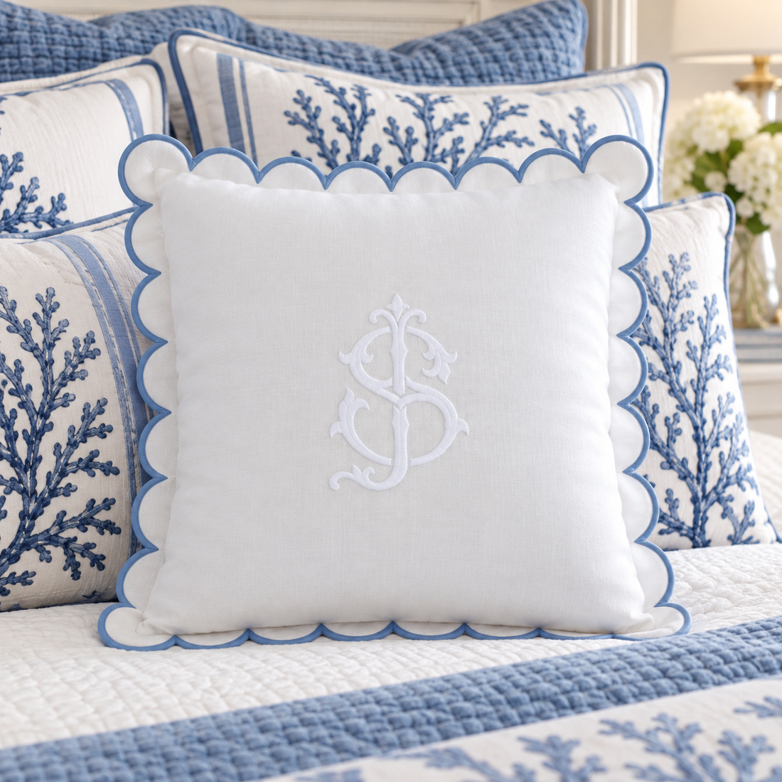 A white decorative throw pillow with a blue scalloped edge and an embroidered monogram, styled on a bed with matching blue and white patterned pillows and bedding.